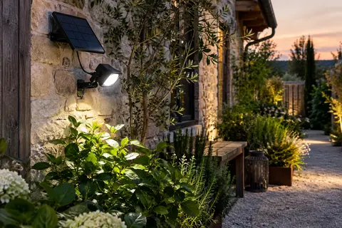 Projecteur solaire noir fixé près d’une allée, avec panneau solaire séparé sur un mur de jardin.